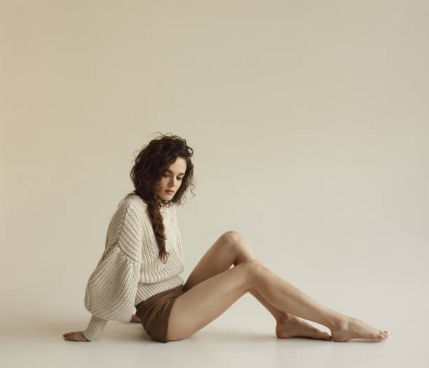 Woman sitting on floor wearing brown shorts and an oversized beige sweater against a neutral background.